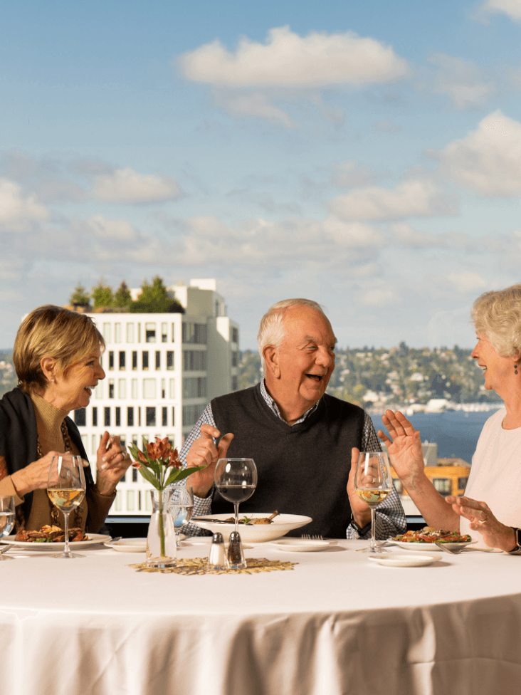 Three friends talk and laugh over dinner with a skyline view.