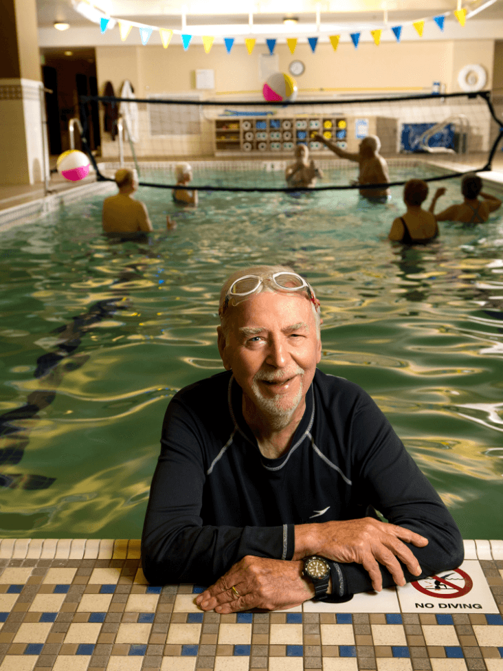 A man smiles in a swimming pool while people play water volleyball in the background.