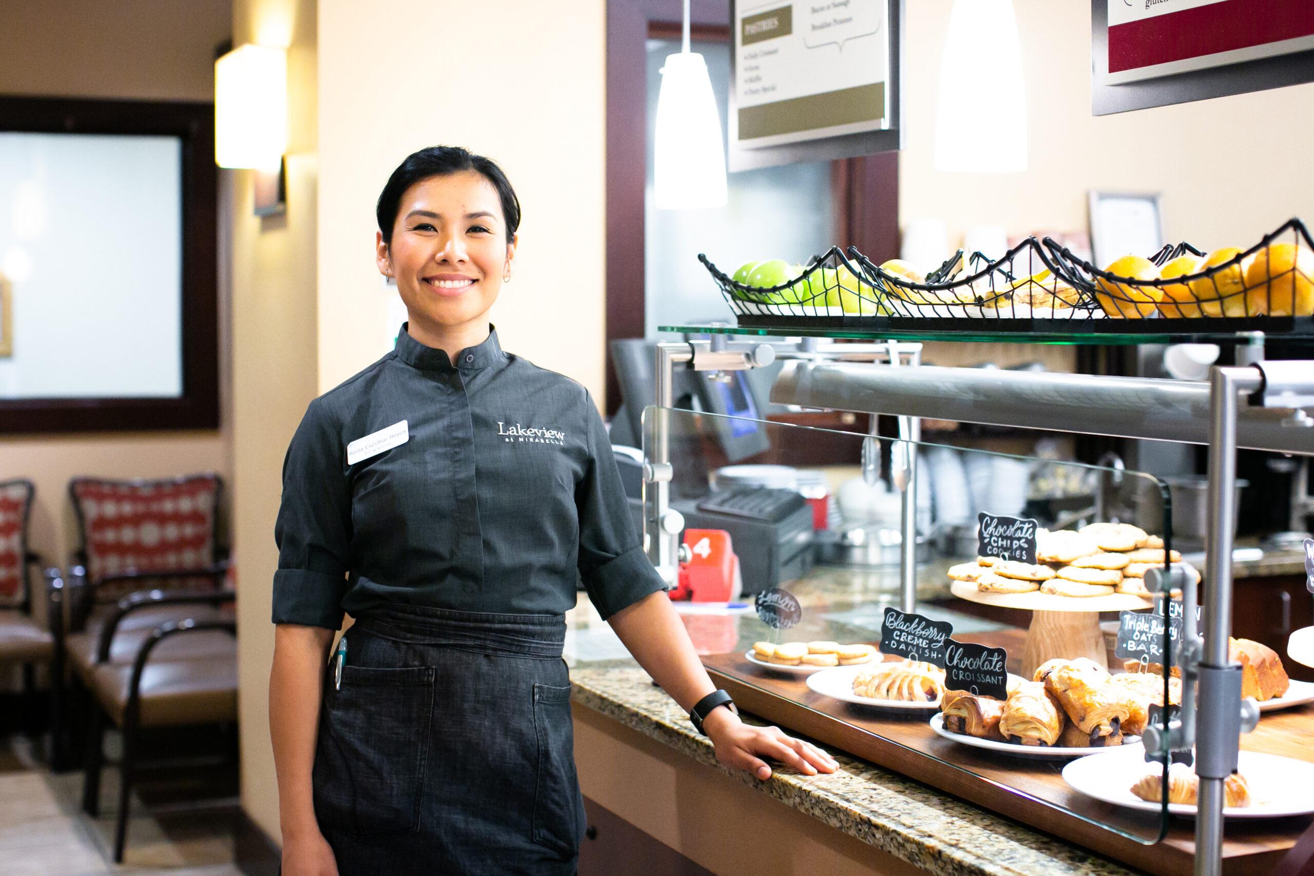 A smiling Bistro Cafe employee stands by a case of pastries.